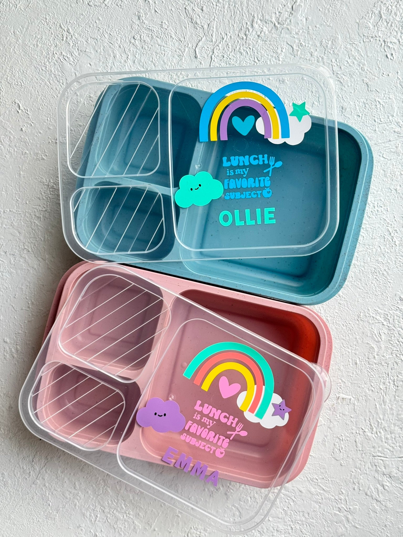 Two bento boxes, one blue and one pink, with rainbow designs and personalized text on a light gray background.