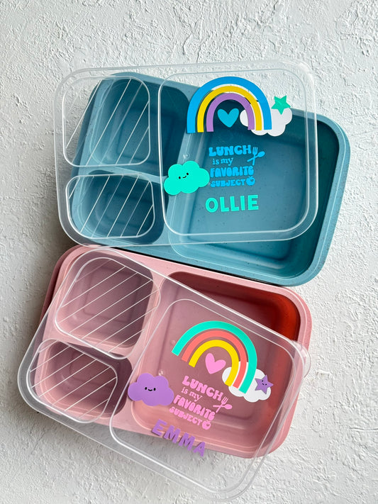 Two bento boxes, one blue and one pink, with rainbow designs and personalized text on a light gray background.